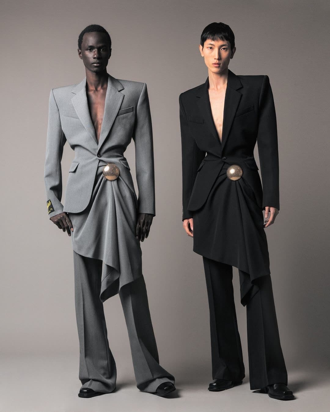 Two menswear looks, sharply tailored grey and black suits with belted waists